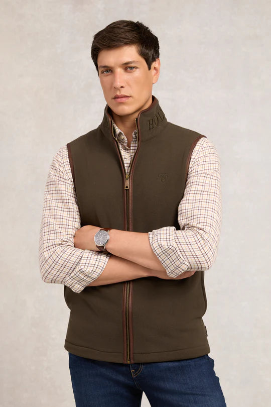 Man wearing a brown vest over a plaid shirt with a neutral background