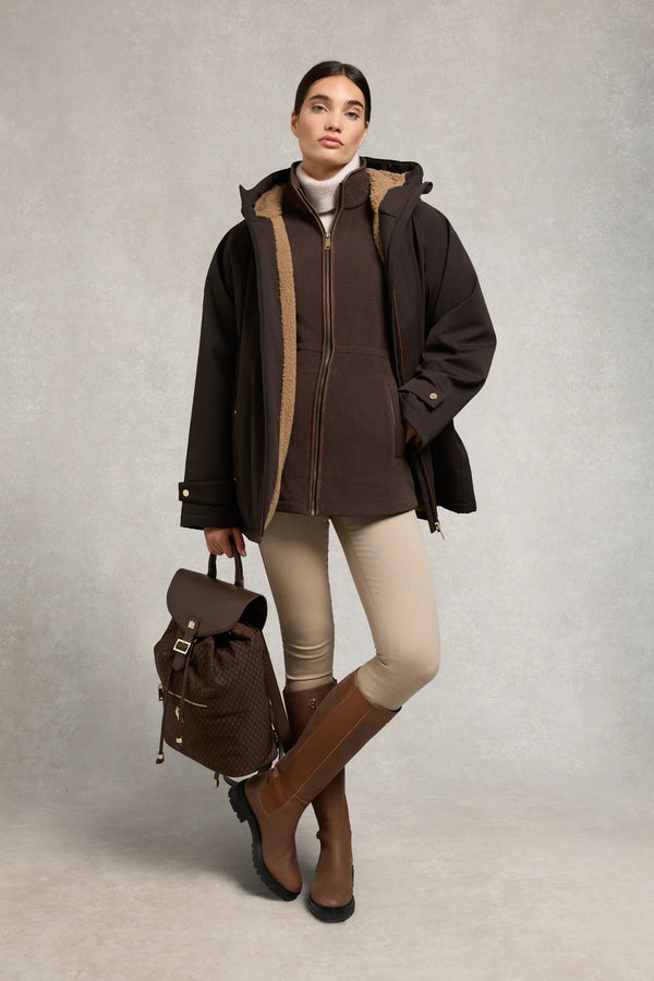 Woman wearing a brown coat, leggings, and boots holding a matching backpack against a neutral background