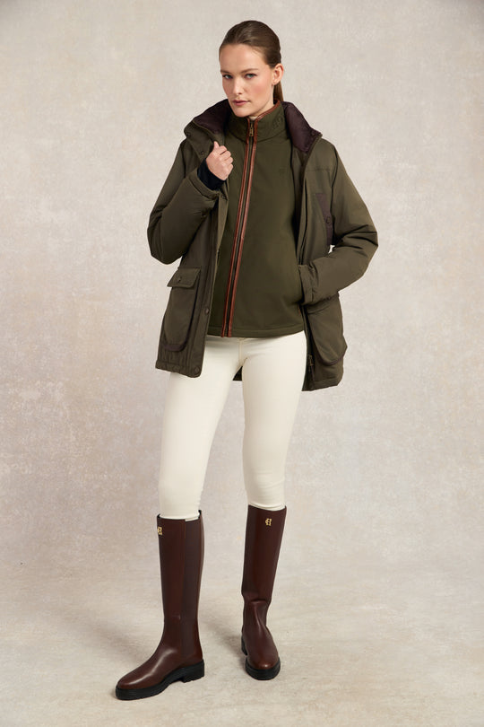 Woman wearing a green jacket, white pants, and brown boots on a neutral background