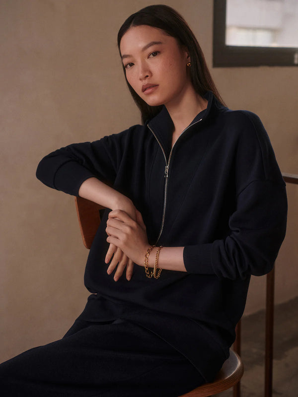 Woman wearing a black zip-up sweatshirt sitting on a chair with a neutral background