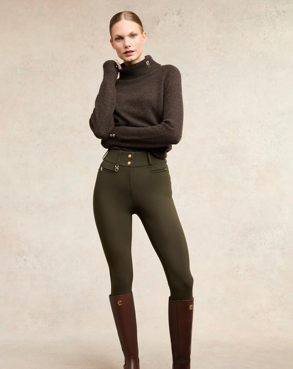 Woman wearing a dark sweater and green riding pants with brown boots on a neutral background