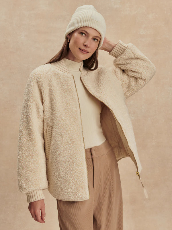 Woman wearing a beige knit cardigan, white sweater, and beige pants against a beige background