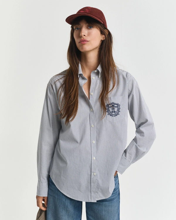 Woman wearing a light gray button-up shirt with a logo, blue jeans, and a maroon cap on a white background