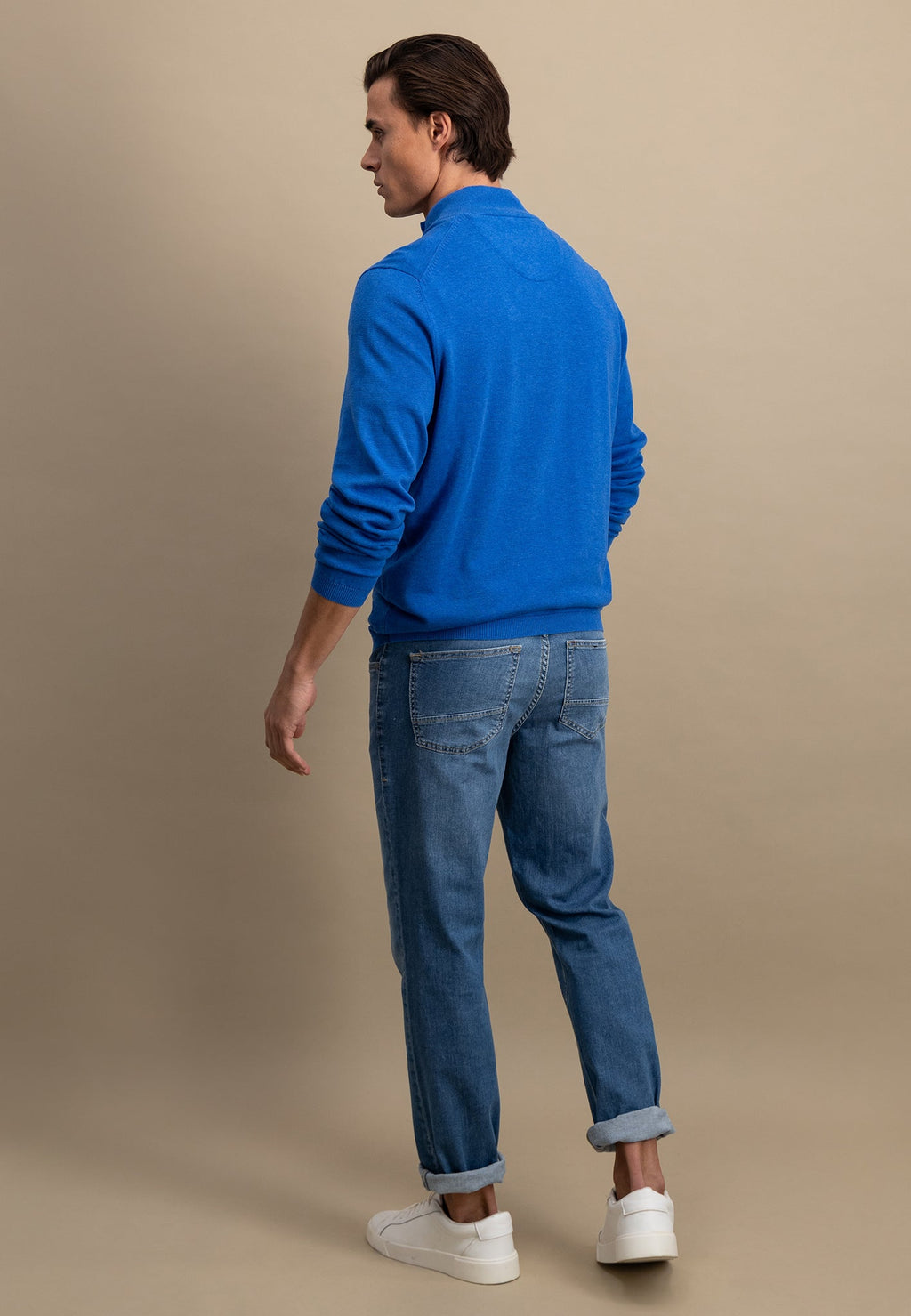 Man wearing a blue sweater and jeans standing against a beige background