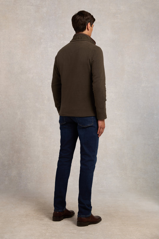 Man wearing a brown jacket and blue jeans against a plain background