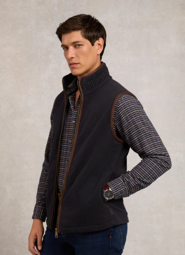 Man wearing a dark vest over a patterned shirt on a neutral background