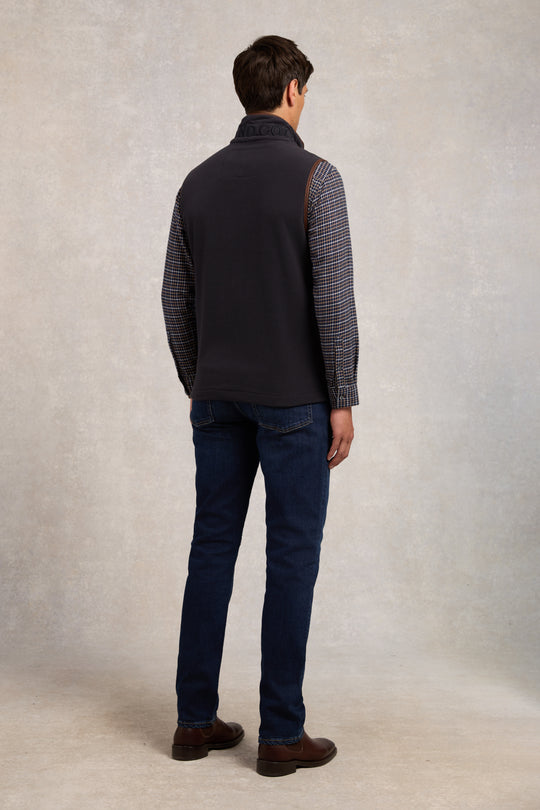 Man wearing a dark vest over a patterned shirt with blue jeans against a neutral background