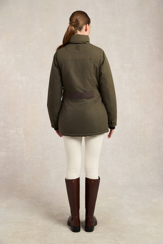 Person wearing an olive green jacket, white pants, and brown boots on a neutral background