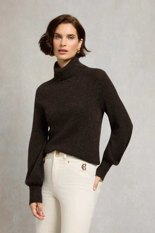Woman wearing a dark brown sweater and white pants against a neutral background