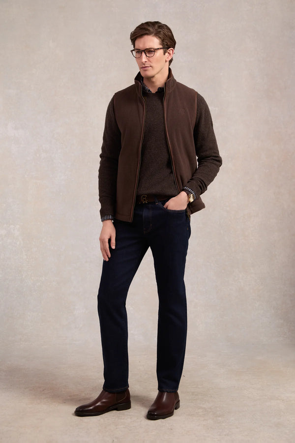 Man wearing a brown jacket, shirt, and jeans standing against a plain background