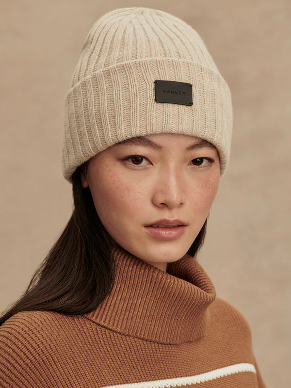 Person wearing a beige knit beanie with a brand logo on a beige background