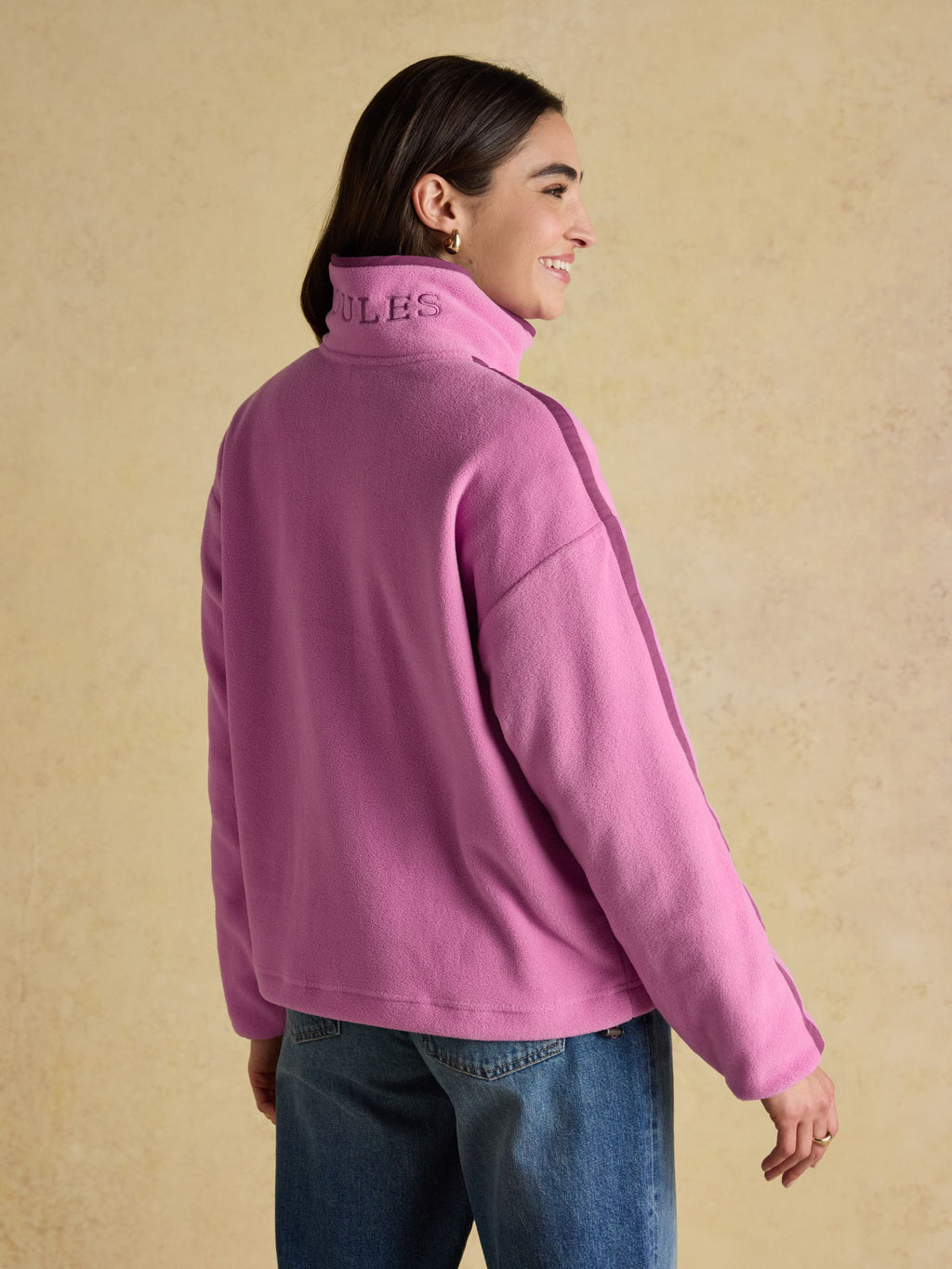 Woman wearing a pink fleece jacket with 'Joules' branding on the back against a beige background