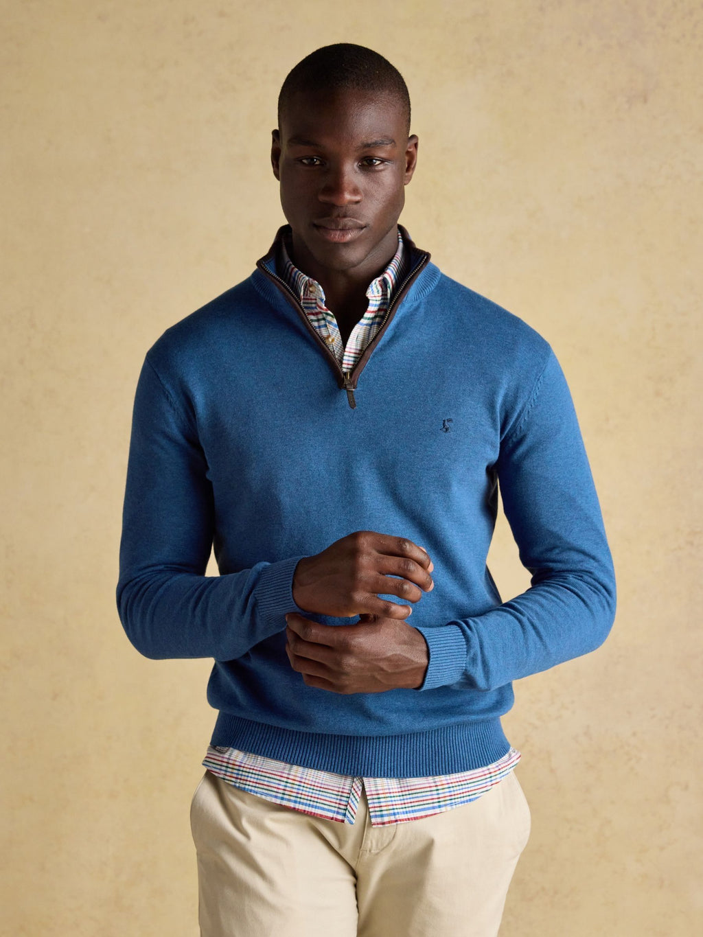 Man wearing a blue sweater over a plaid shirt with beige pants against a beige background