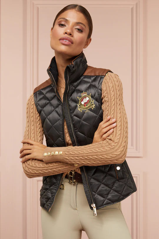 Woman wearing a black quilted vest with a brand logo against a beige wall.