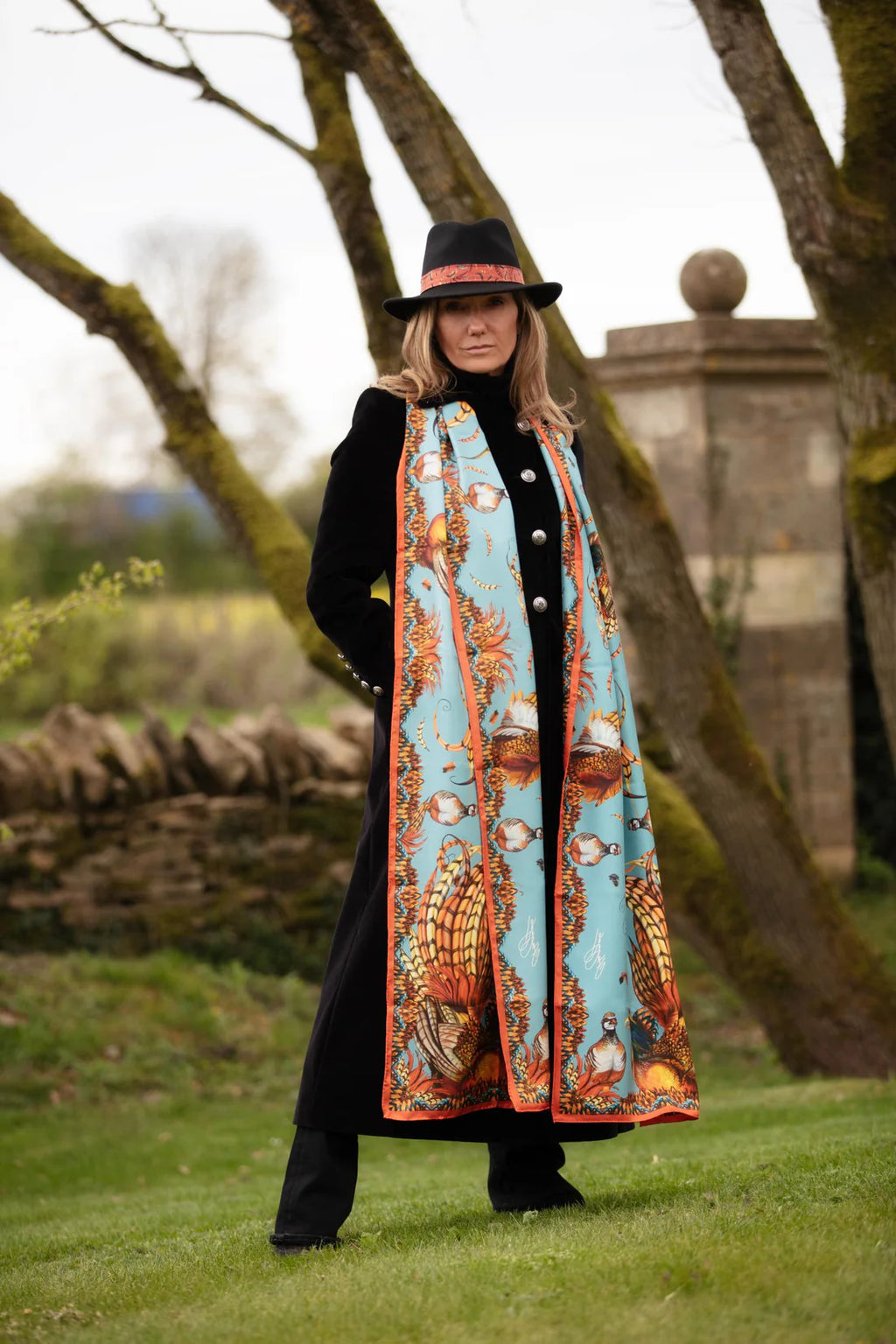 Woman wearing a long, patterned scarf outdoors with trees and a stone wall in the background.