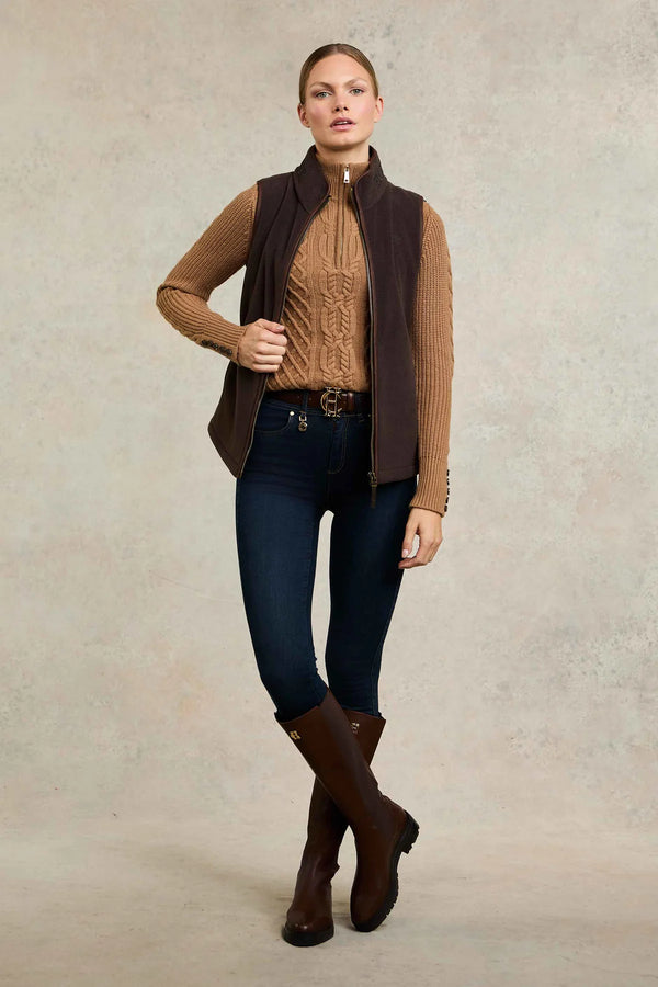 Woman wearing a brown vest, sweater, and jeans with boots on a plain background