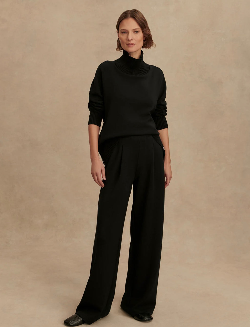 Woman wearing a black turtleneck sweater and wide-leg pants against a beige background