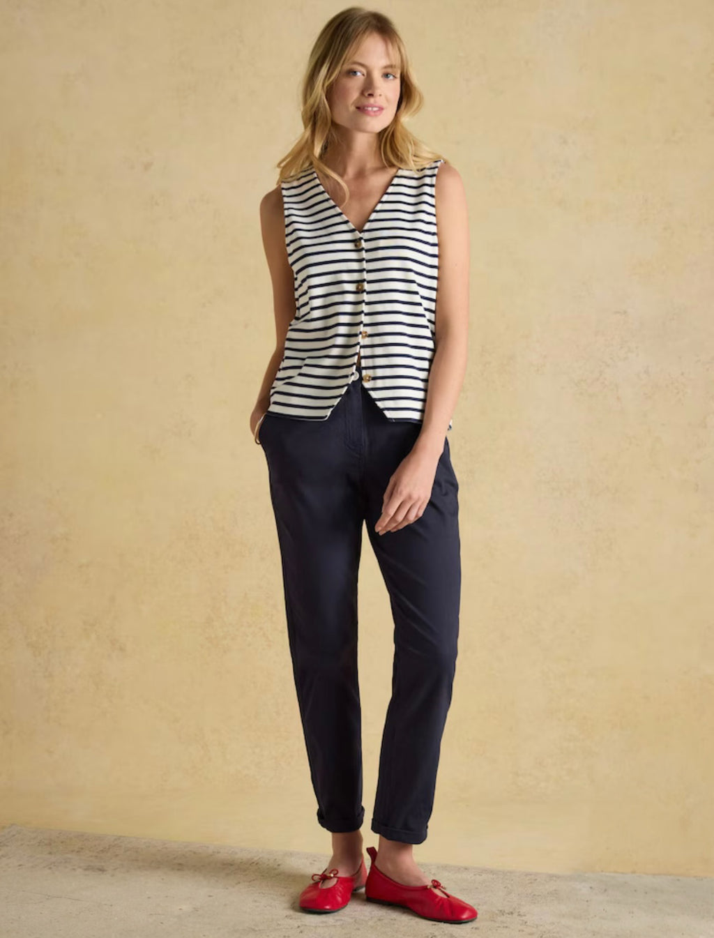 Woman wearing a striped sleeveless top and navy pants with red shoes against a beige background