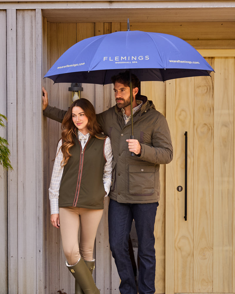 Two people standing under a blue umbrella with 'Flemings' branding, in front of a wooden door.