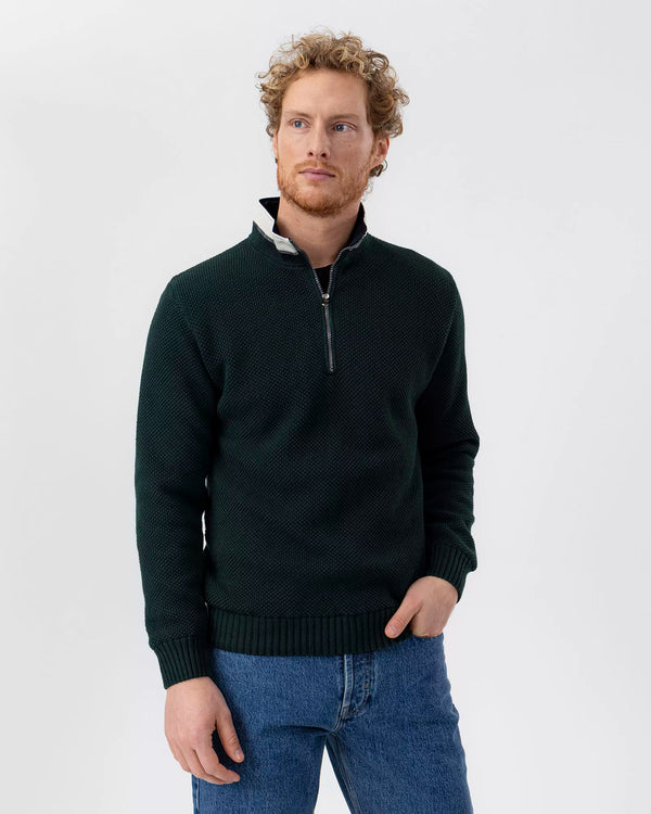 Man wearing a green quarter-zip sweater and blue jeans on a white background