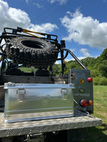 Back of a truck with a large metal box and tire on a grassy area with trees and blue sky.