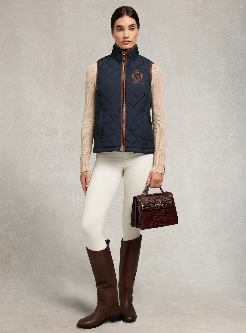 Woman wearing a navy quilted vest, white pants, and brown riding boots holding a brown bag on a gray background.