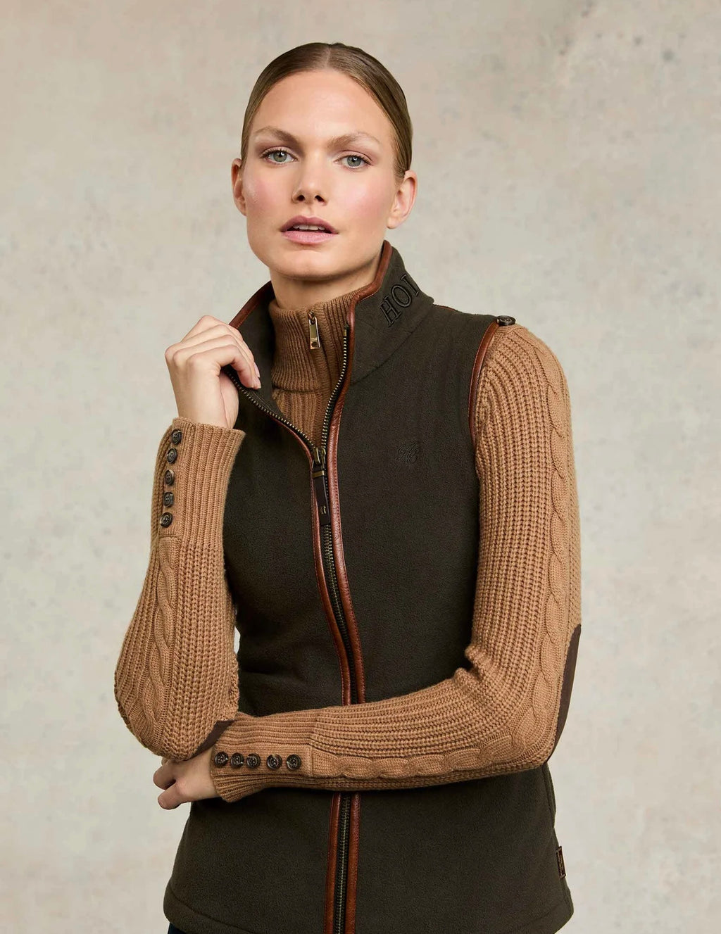 Woman wearing a brown knit sleeveless jacket over a black outfit against a neutral background