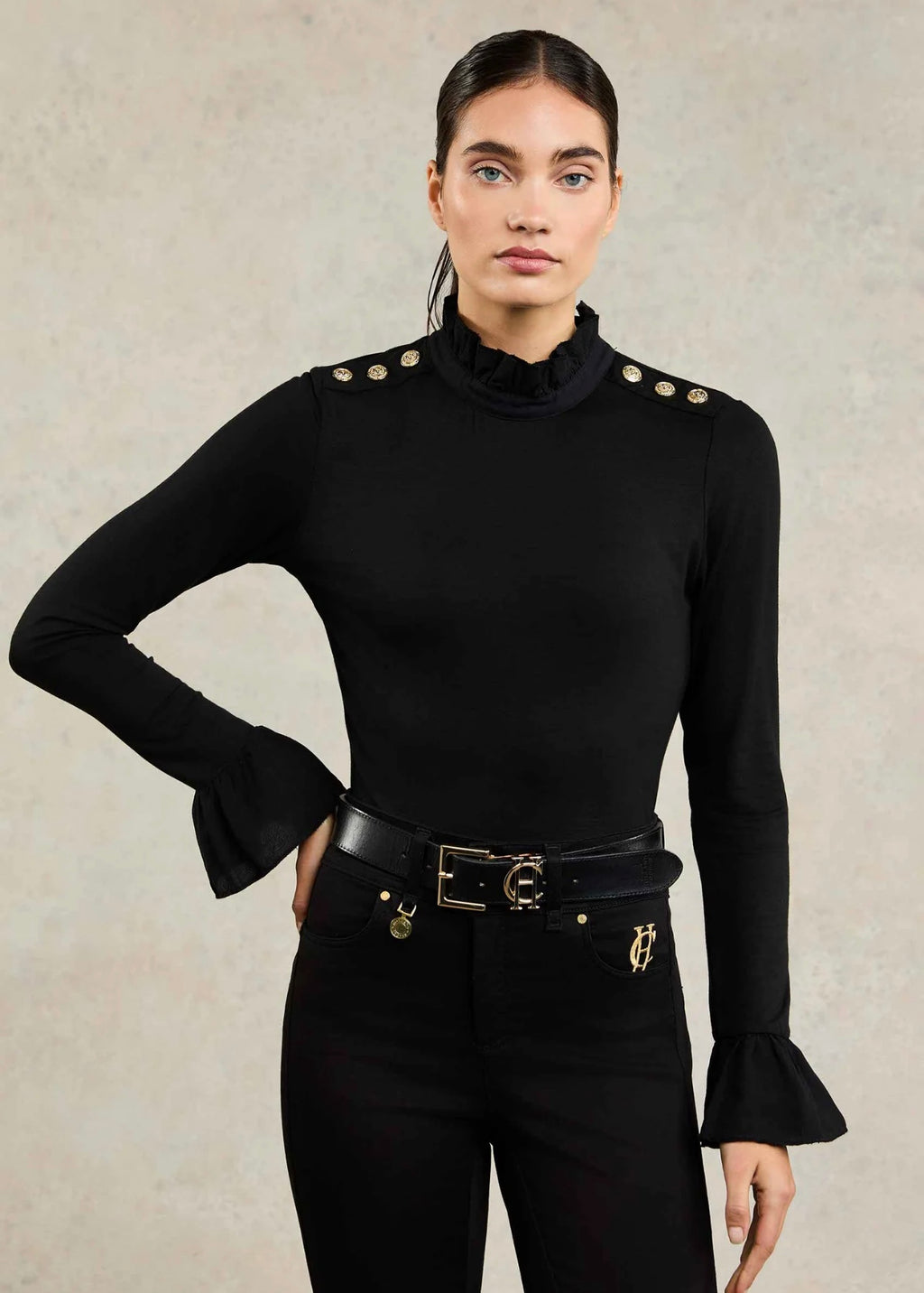 Woman wearing a black long-sleeve top with gold buttons and black pants against a beige background