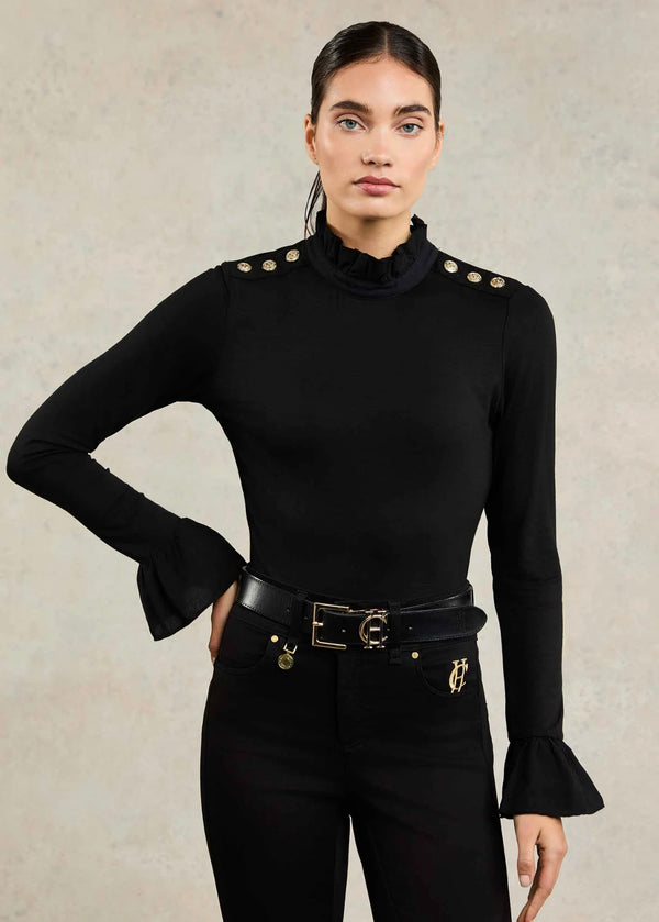 Woman wearing a black long-sleeve top with gold buttons and black pants against a beige background