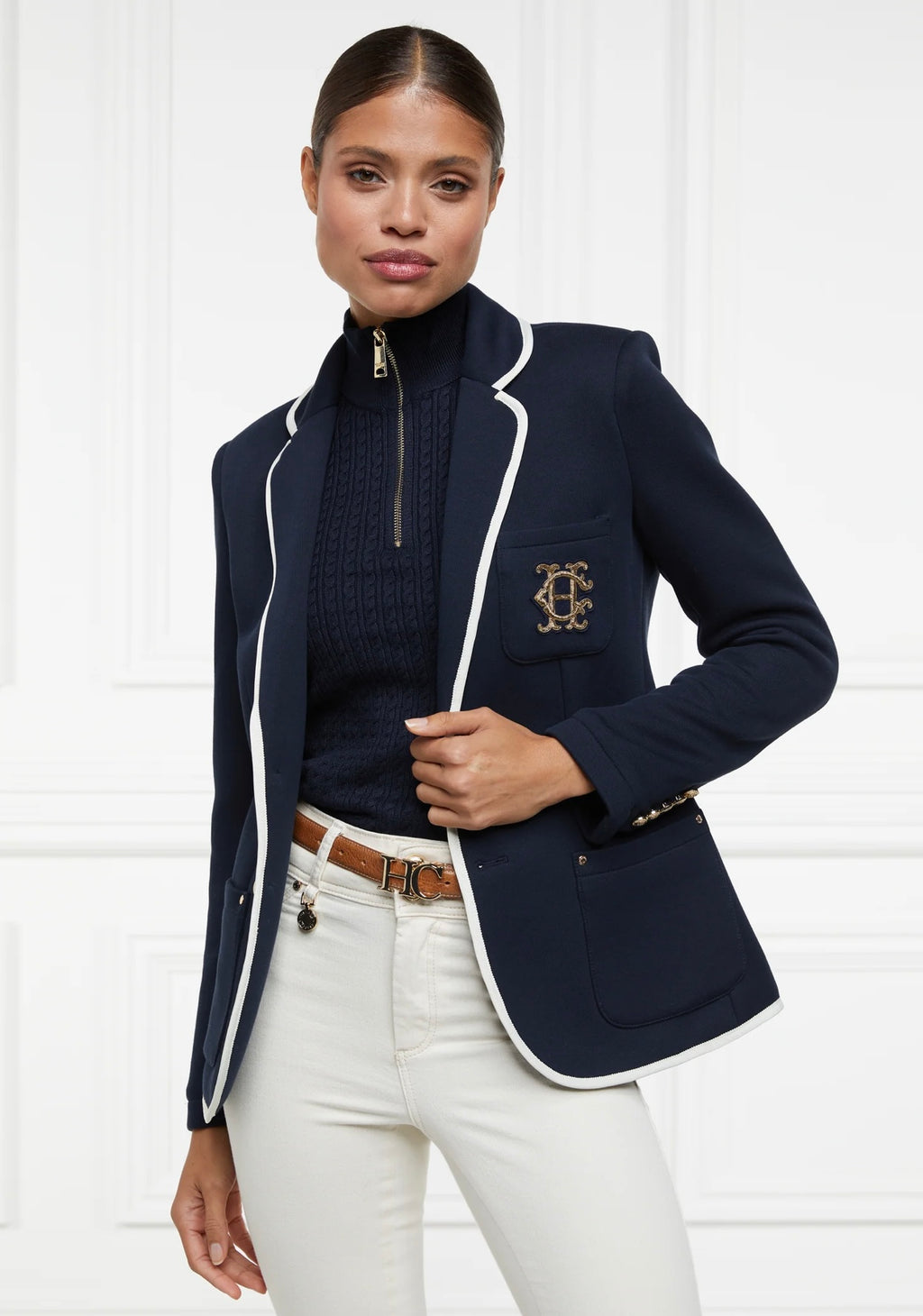 Woman wearing a navy blazer with white trim and a logo, white pants, and a dark sweater.