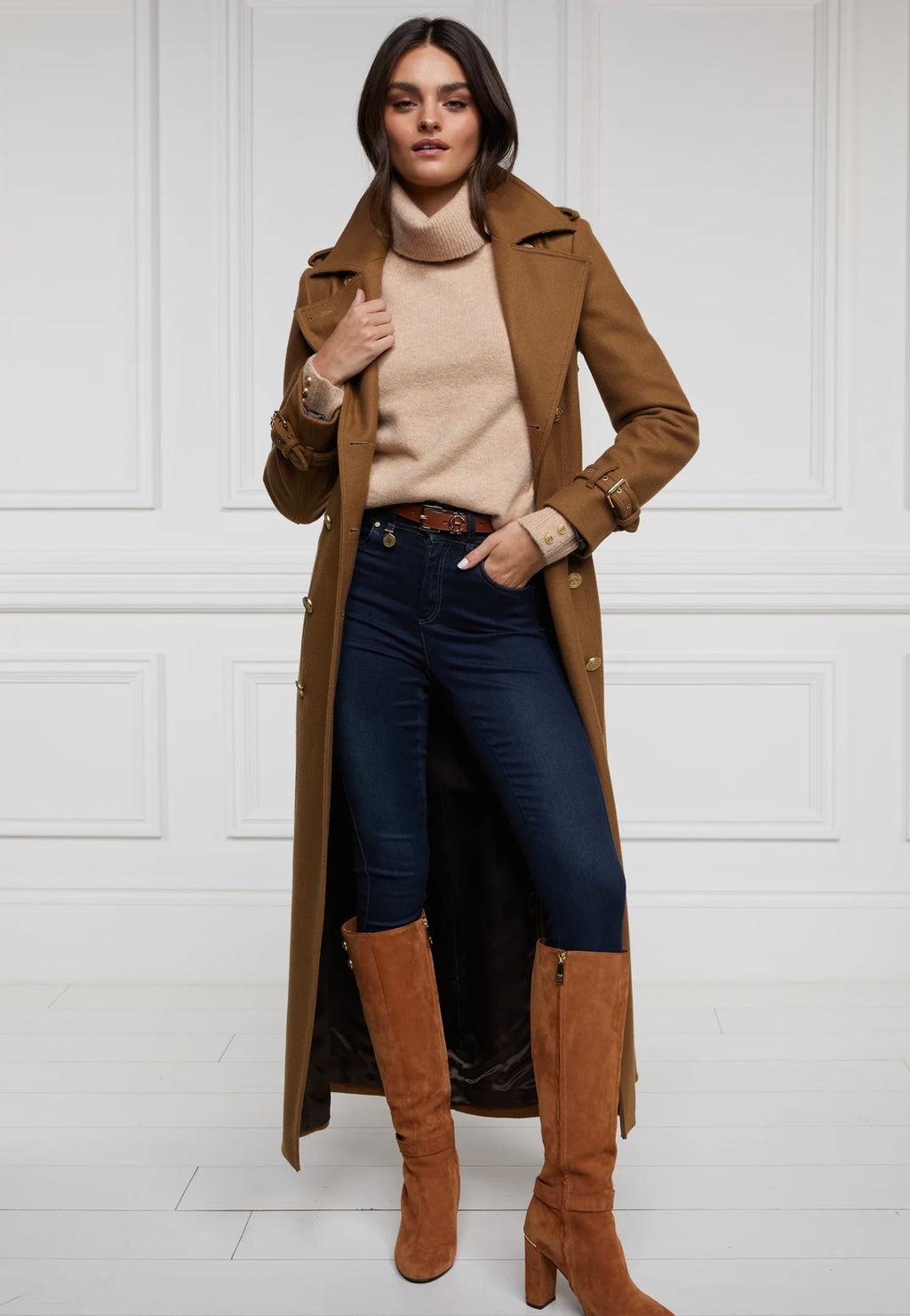 Woman wearing a long brown coat, beige sweater, dark jeans, and brown knee-high boots against a white background.