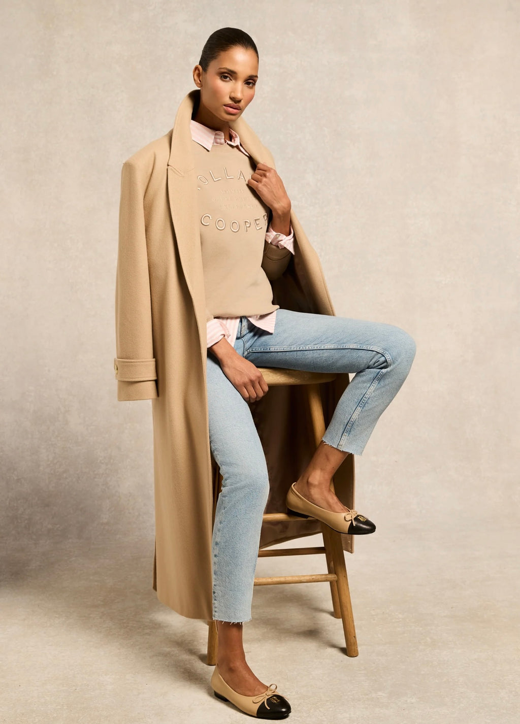 Woman sitting on a stool wearing a beige coat, light blue jeans, and a beige sweater with 'COCO' text.