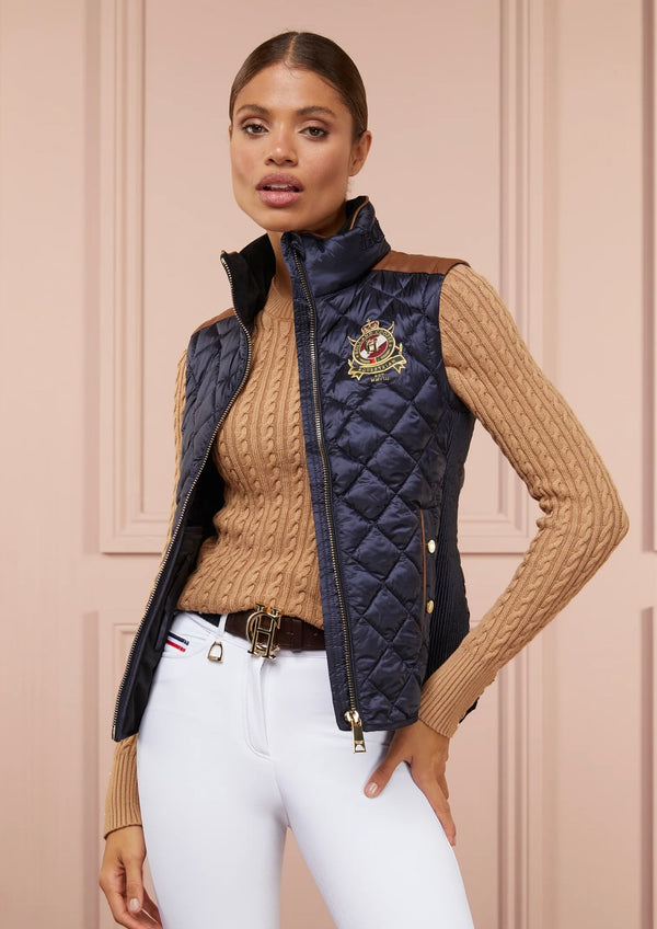 Woman wearing a navy quilted vest with a brand logo, beige sweater, and white pants against a beige wall.