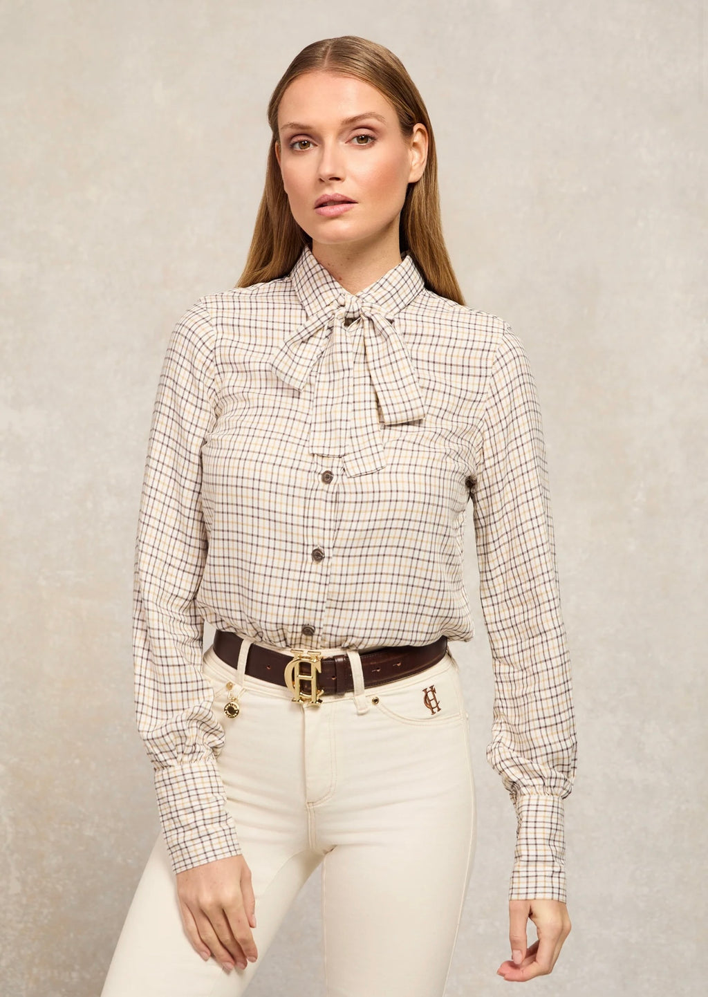Woman wearing a checkered blouse with a bow tie and white pants against a neutral background