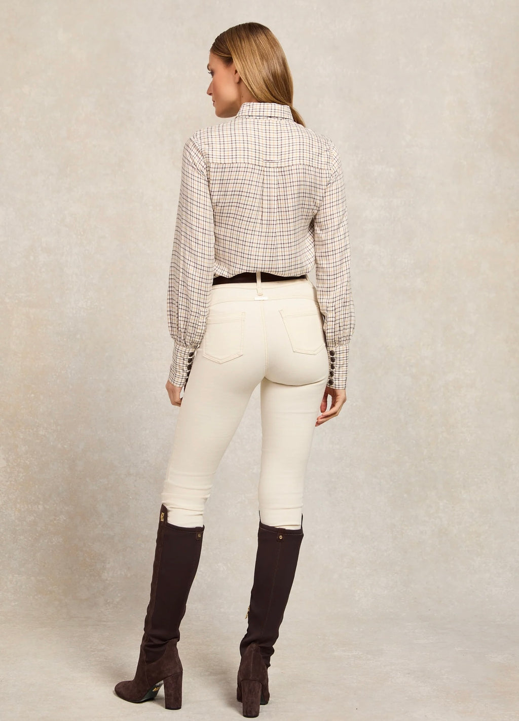 Woman wearing a checkered shirt, white pants, and black knee-high boots on a plain background