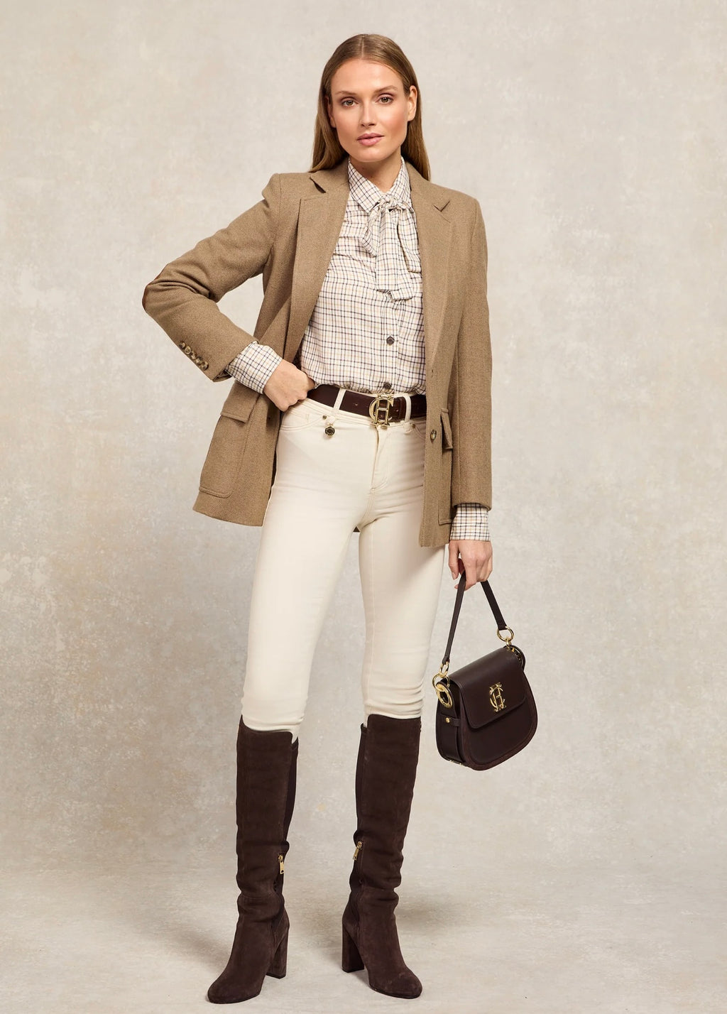 Woman wearing a brown blazer, white shirt, white pants, and brown knee-high boots, holding a black handbag.