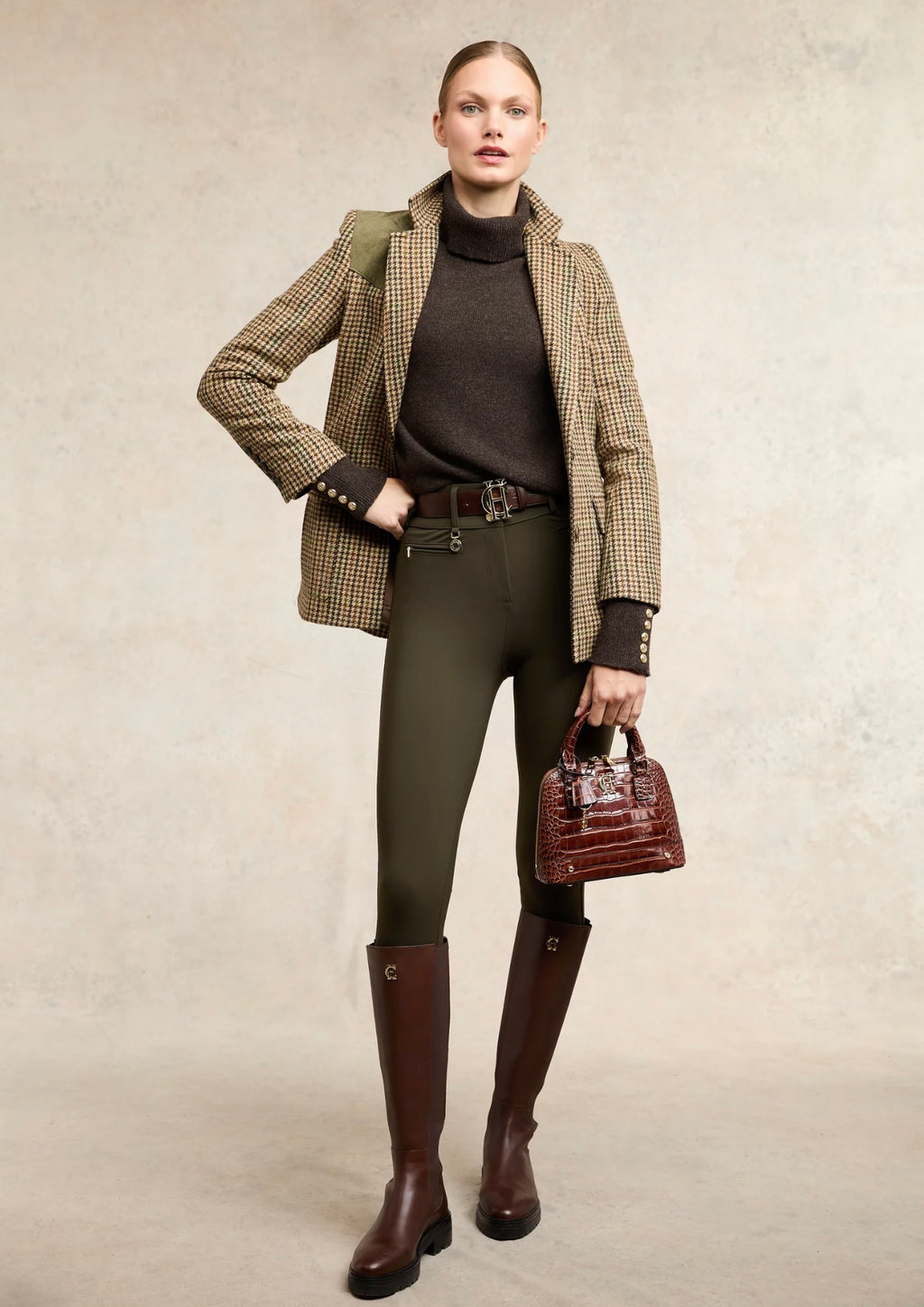 Woman in equestrian attire with a plaid blazer, green leggings, brown boots, and a brown handbag on a neutral background.