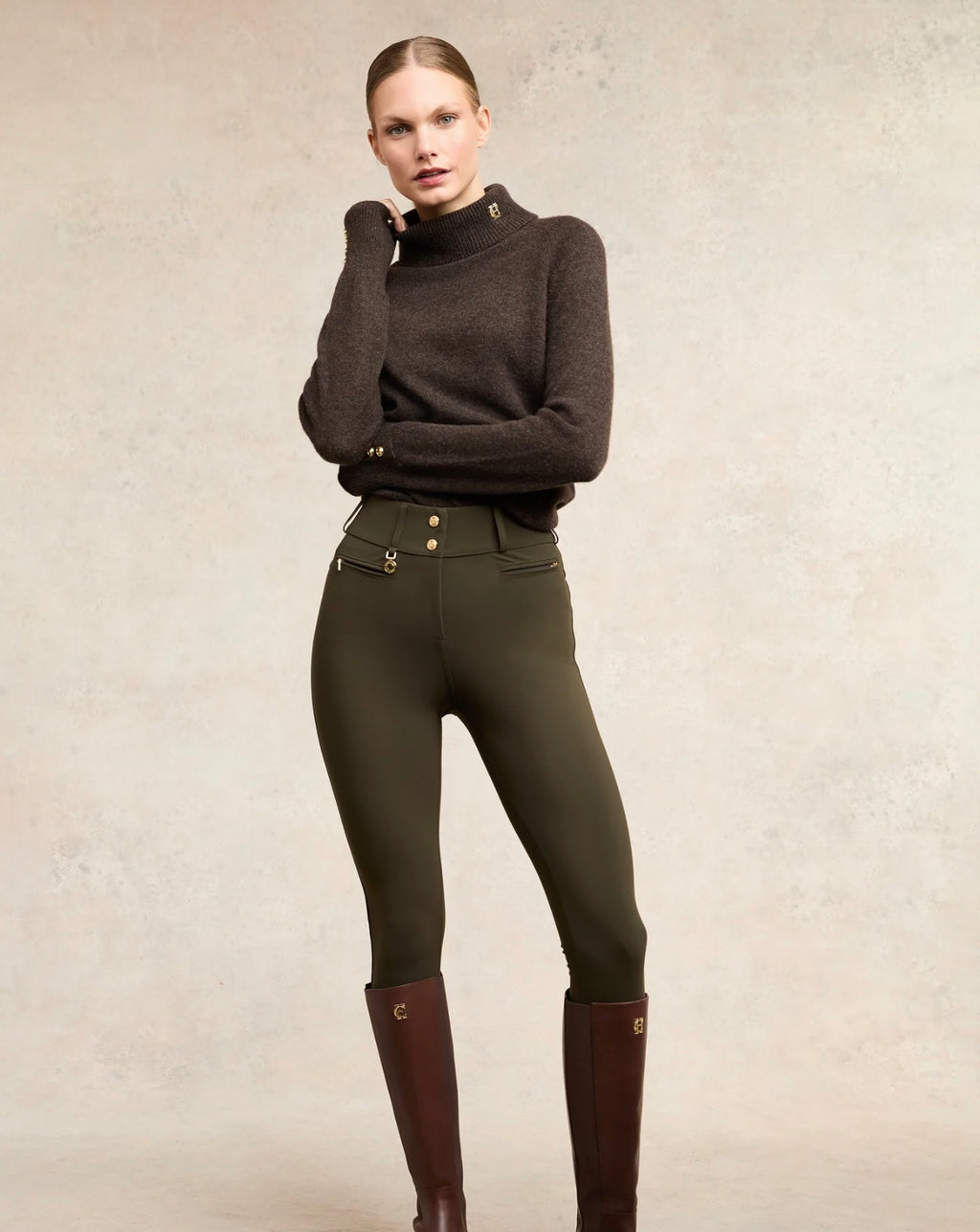 Woman wearing a dark sweater and green riding pants with brown boots on a neutral background