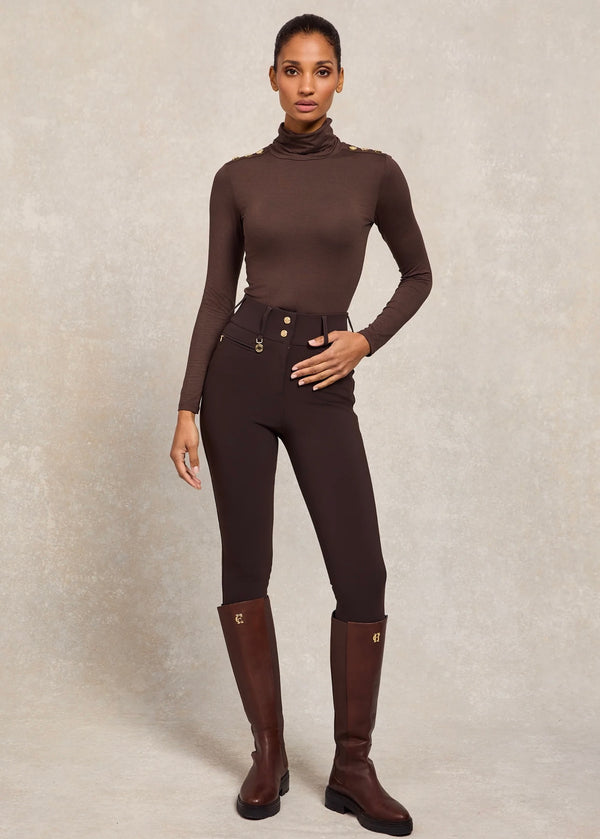 Woman wearing a brown equestrian outfit with leggings and boots on a neutral background