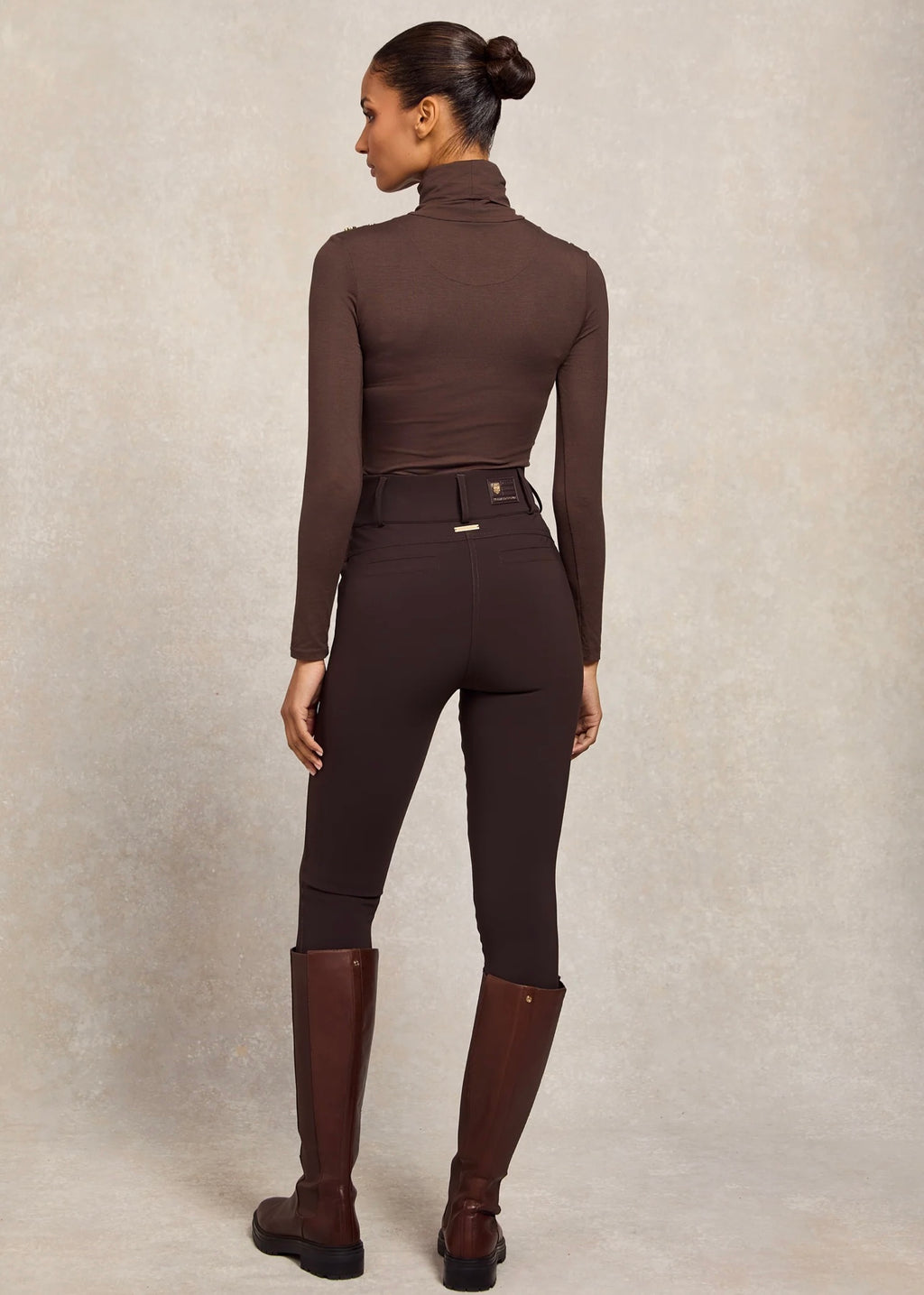 Person wearing a brown equestrian jumpsuit with tall boots on a neutral background