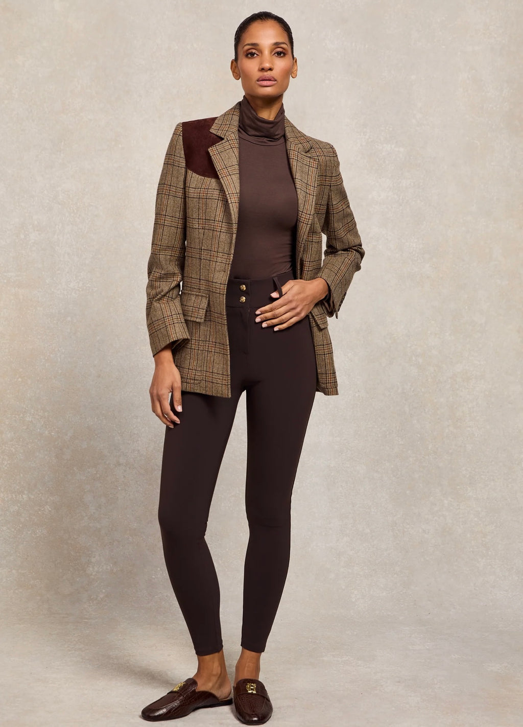 Woman wearing a brown checkered blazer over a brown turtleneck and black leggings on a neutral background