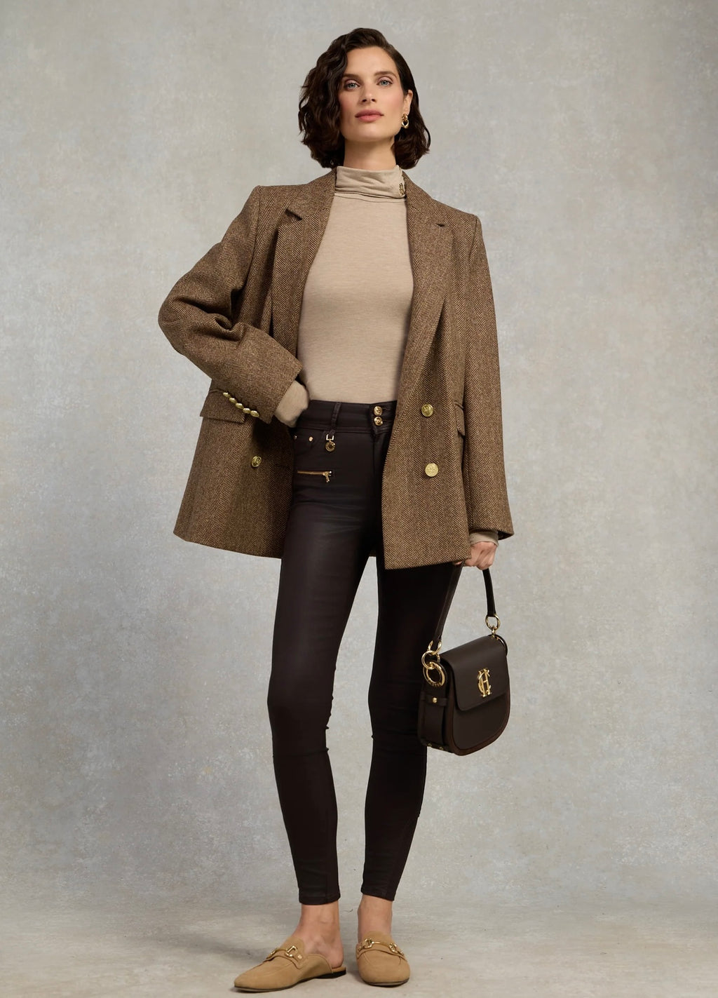 Woman wearing a brown coat, beige sweater, black pants, and holding a handbag against a plain background