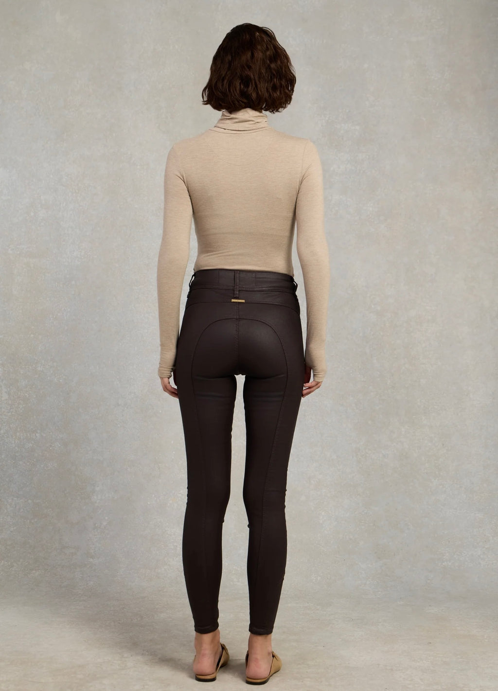 Person wearing a beige turtleneck and dark brown riding pants against a gray background