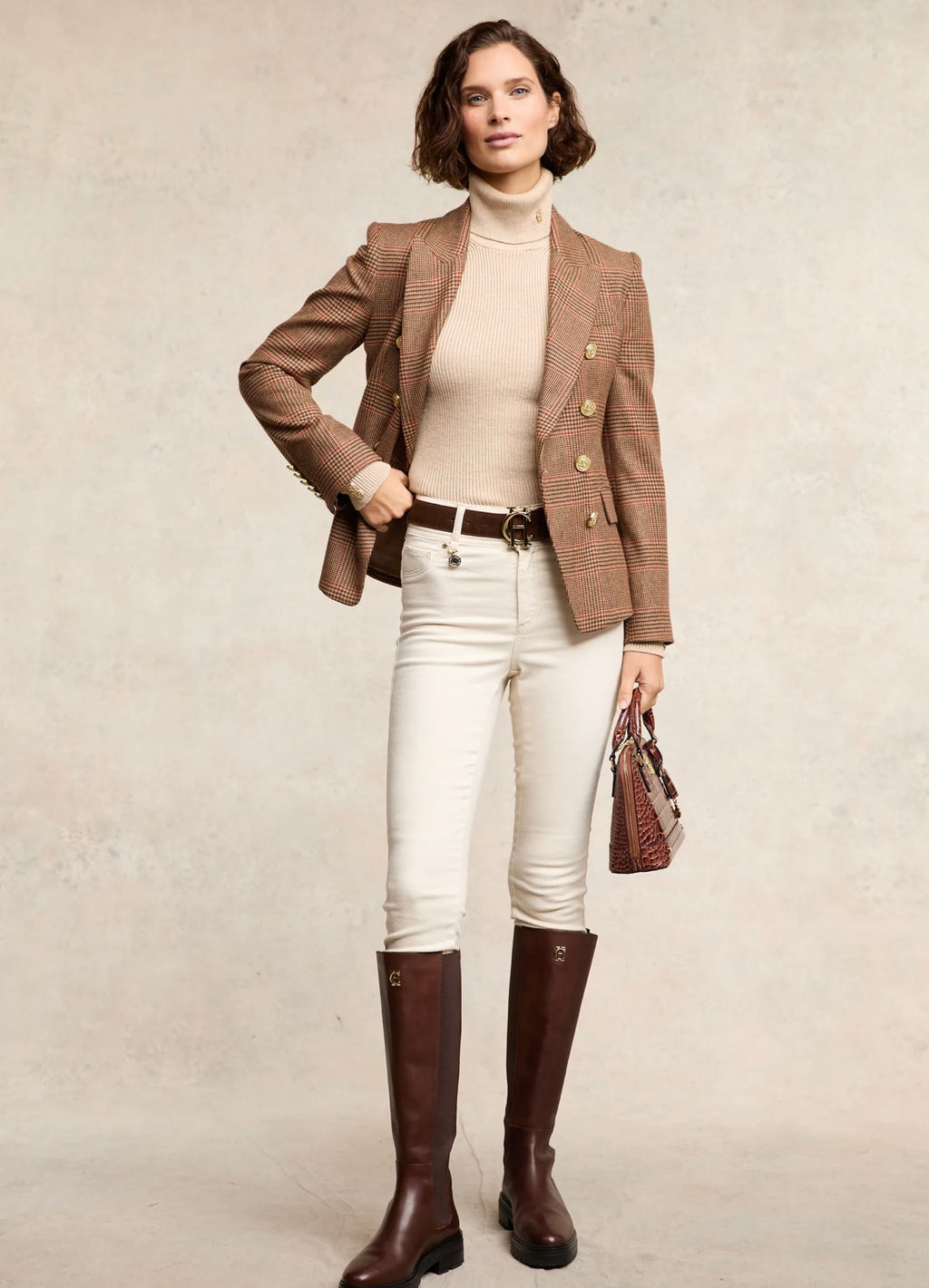 Woman wearing a brown blazer, white turtleneck, white pants, and brown riding boots against a neutral background.