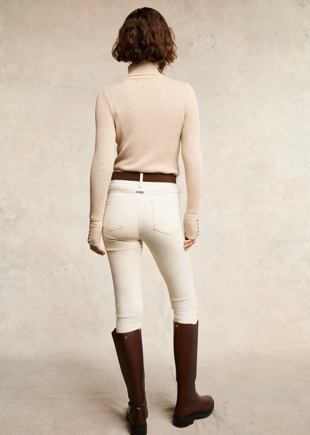 Person wearing a beige long-sleeve shirt, white pants, and brown riding boots on a plain background