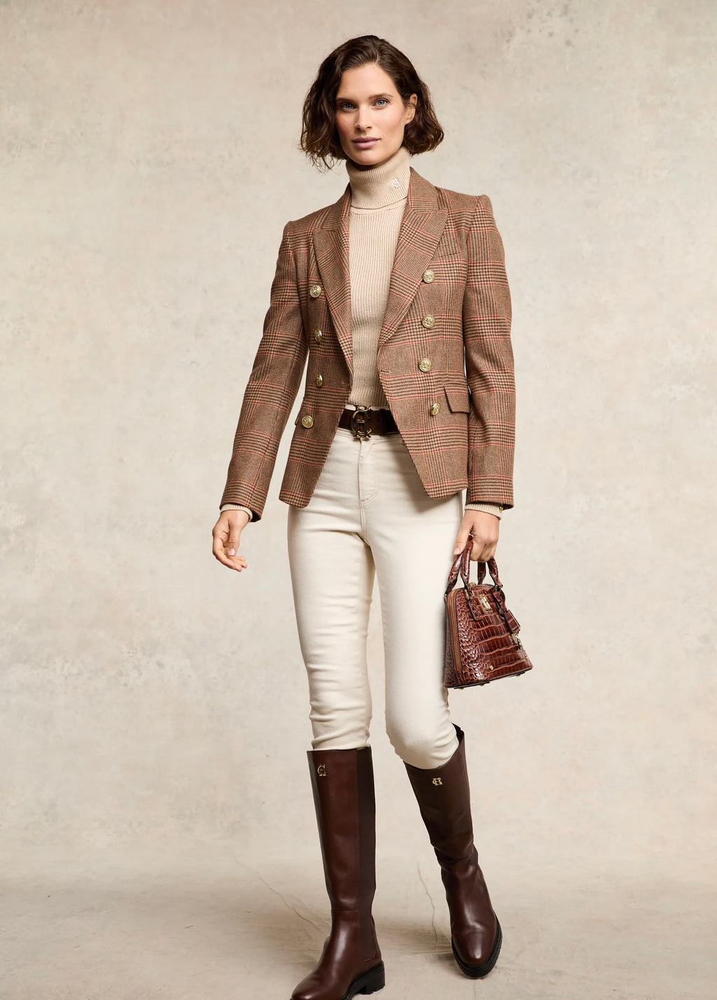 Woman wearing a brown checkered blazer, white pants, and brown knee-high boots holding a handbag.