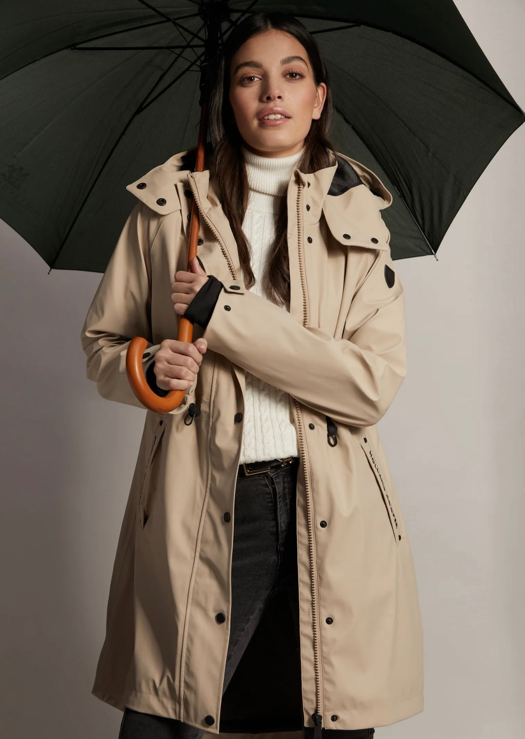 Woman wearing a beige trench coat holding a black umbrella against a plain background