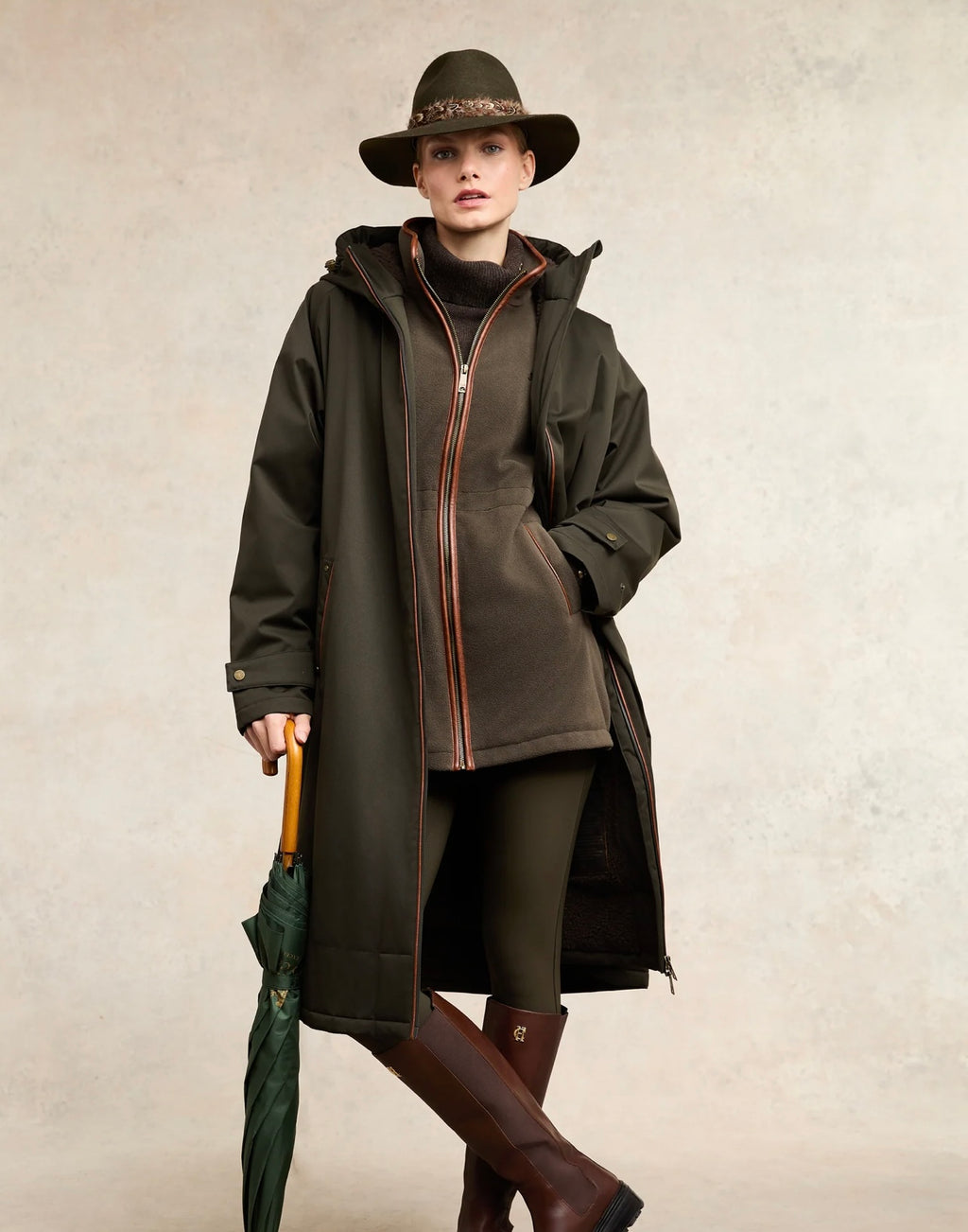 Person wearing a long dark coat, hat, and boots holding an umbrella on a neutral background