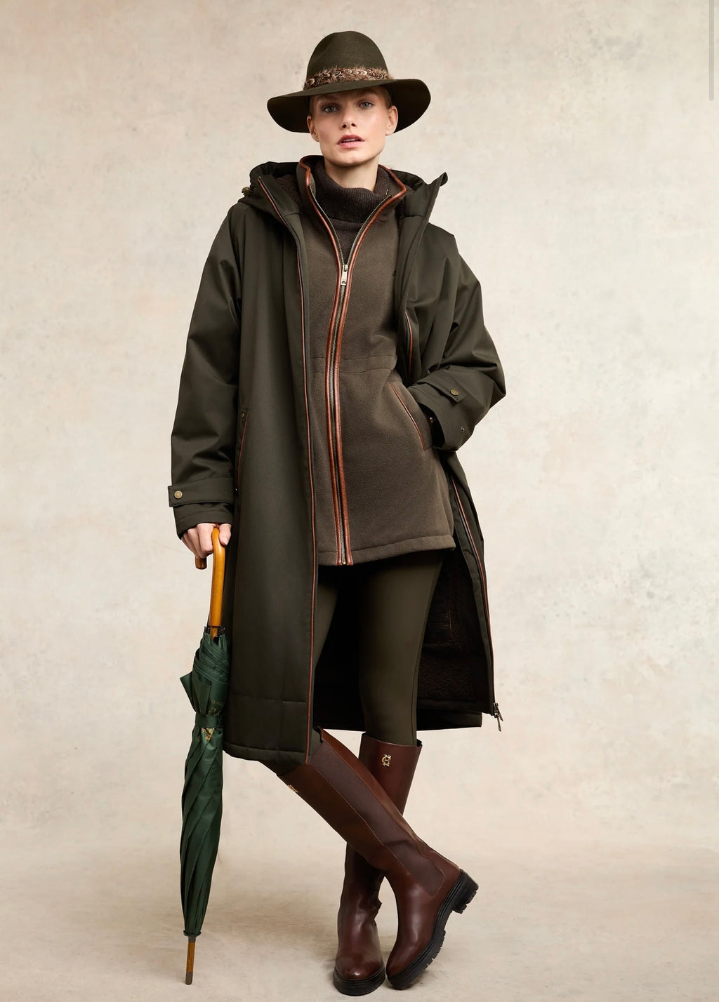 Woman wearing a long dark coat, hat, and riding boots holding an umbrella on a neutral background