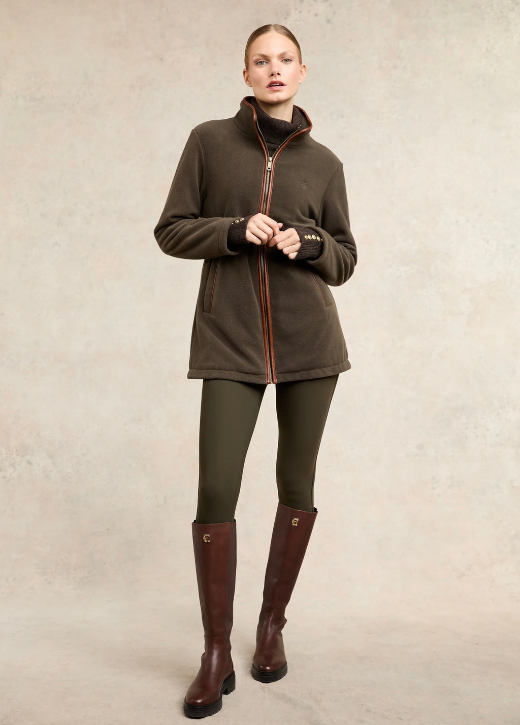 Woman wearing a brown jacket, green leggings, and brown riding boots on a beige background
